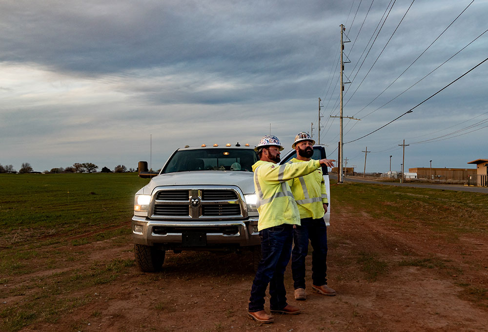 Dos trabajadores de servicios públicos, Kevin Walker y Dylan Edgmon, con camisetas de seguridad amarillas y cascos, están de pie junto a un camión blanco al atardecer; uno de ellos sostiene un portapapeles y señala hacia la distancia.